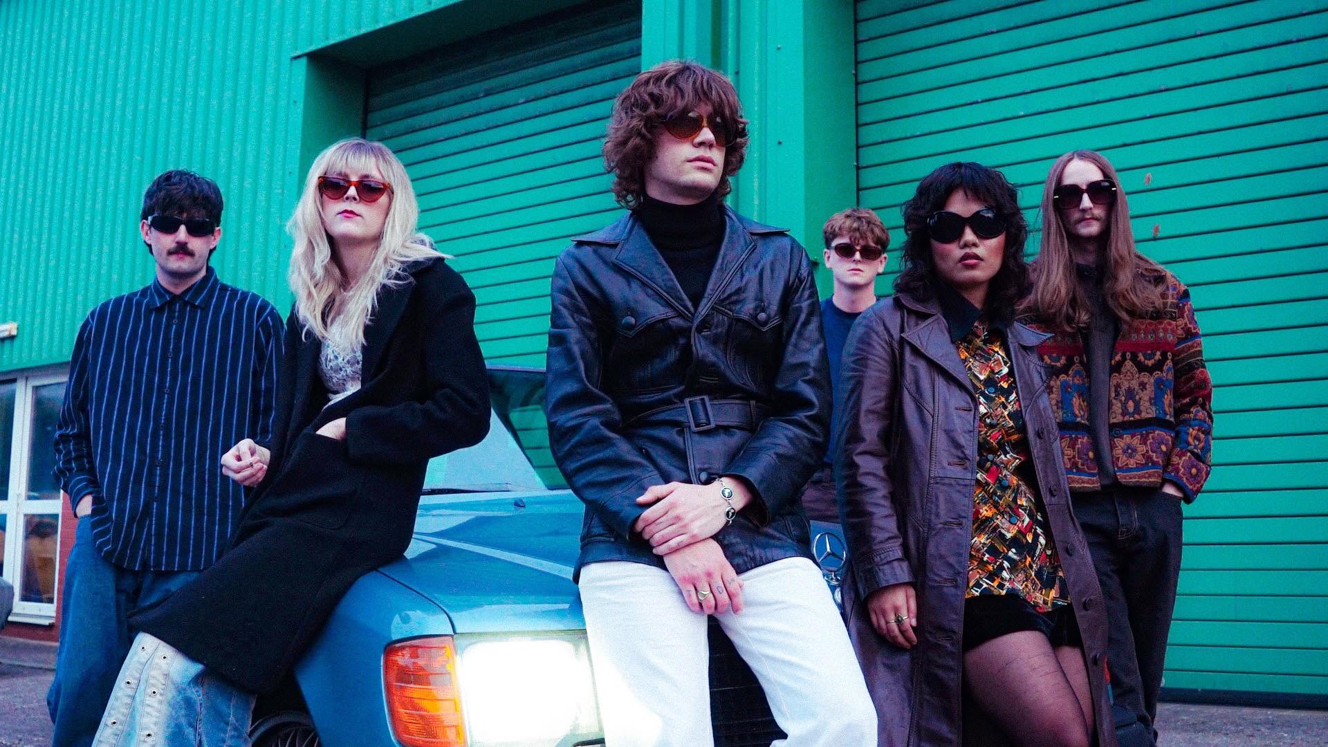 Five band members in retro sunglasses and vintage clothing pose before a green warehouse and a blue car, The Unsigned Guide